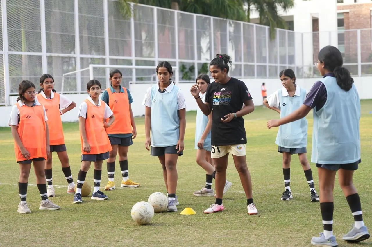 Aditi Chauhan coaching a group of schoolgirls in bibs with footballs on a grass pitch