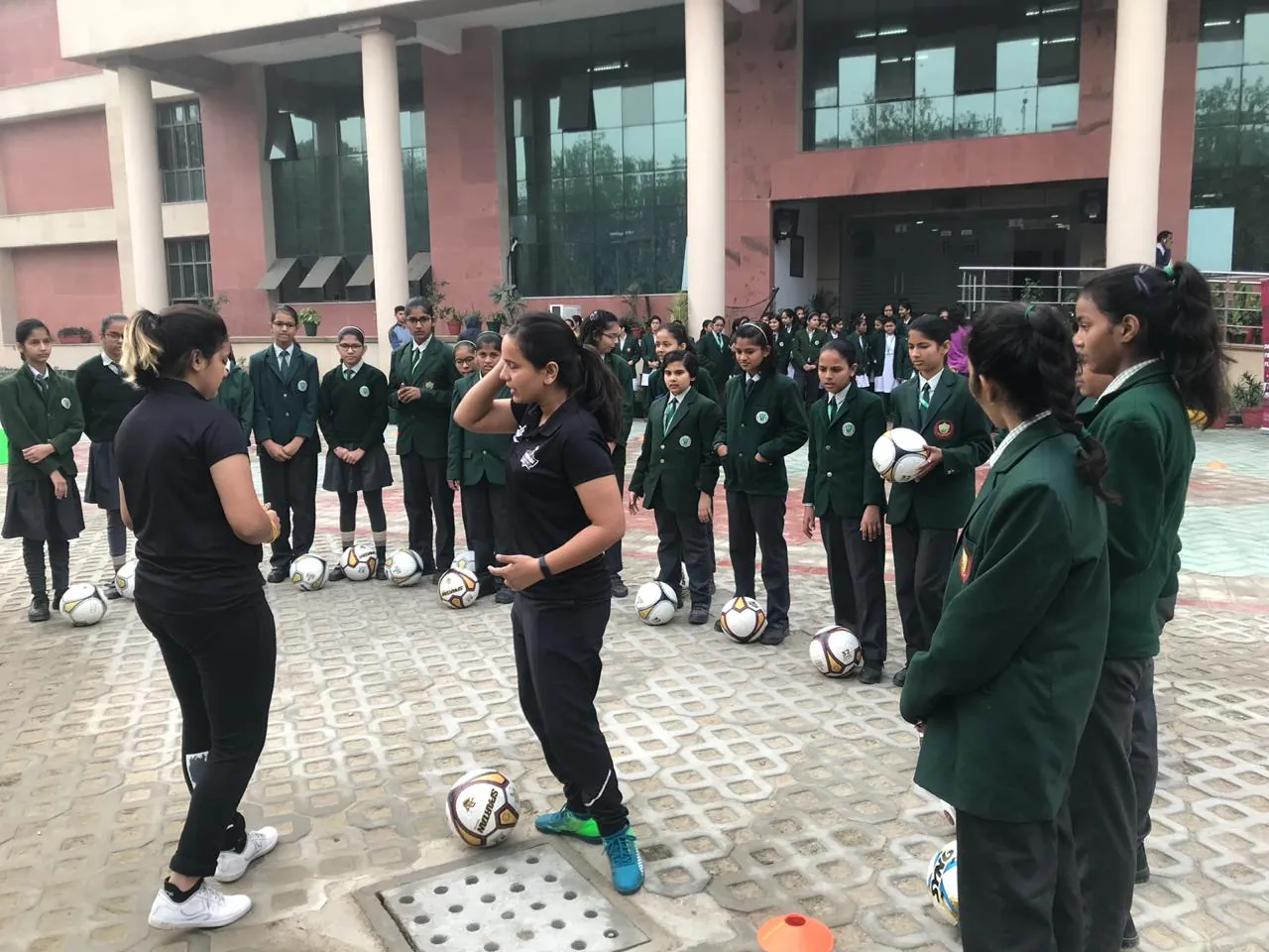 A She Kicks coach demonstrating ball control to a circle of schoolgirls in green blazers at a school