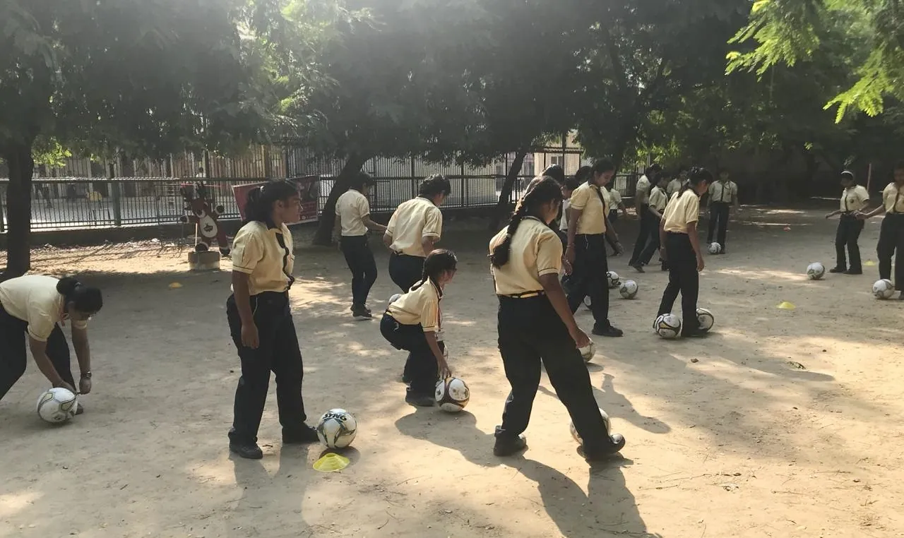 Schoolgirls in uniform dribbling footballs across a dusty ground at a grassroots session