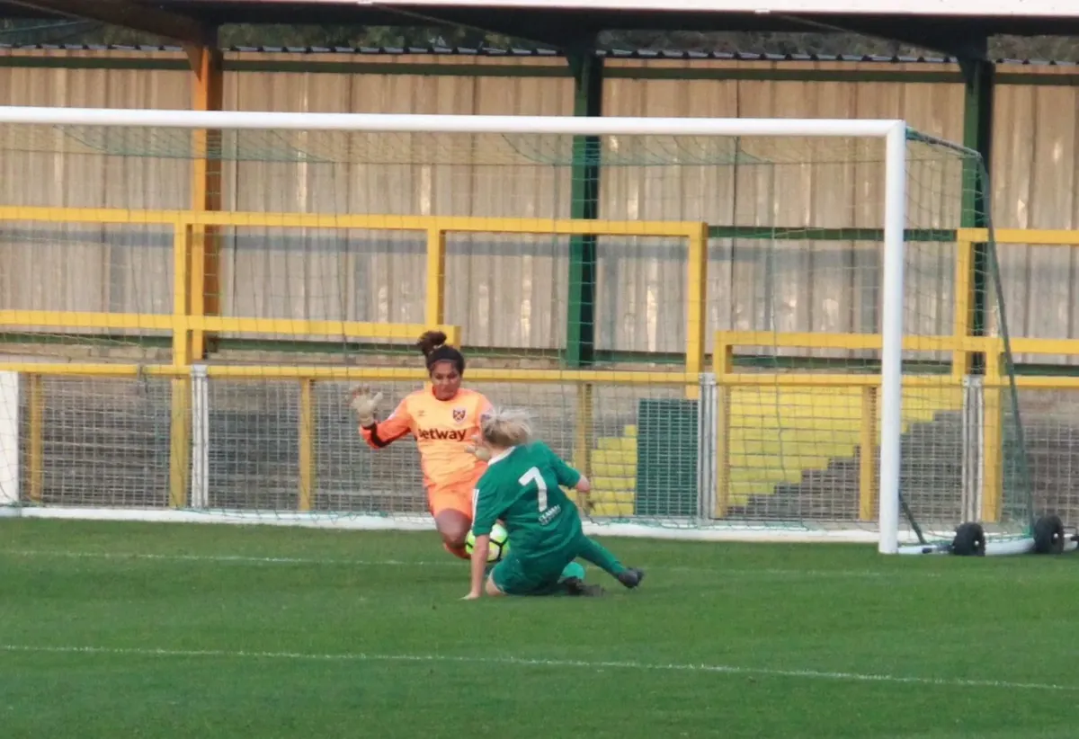 Aditi Chauhan in her orange West Ham United goalkeeper kit making a one-on-one save against an attacker.