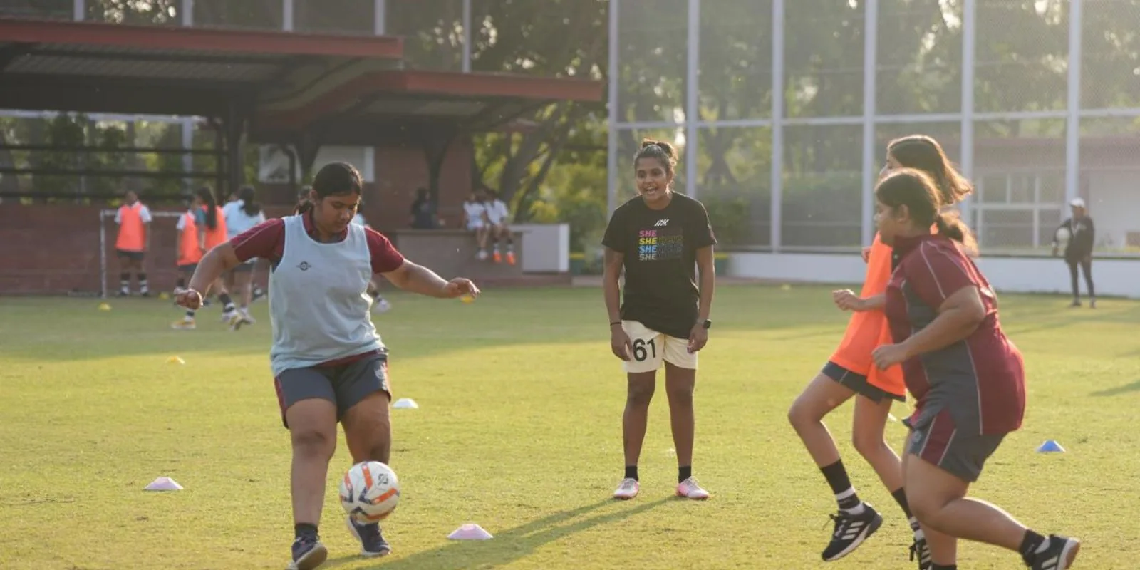 Aditi Chauhan coaching two girls in bibs through a one-on-one drill on a grass pitch