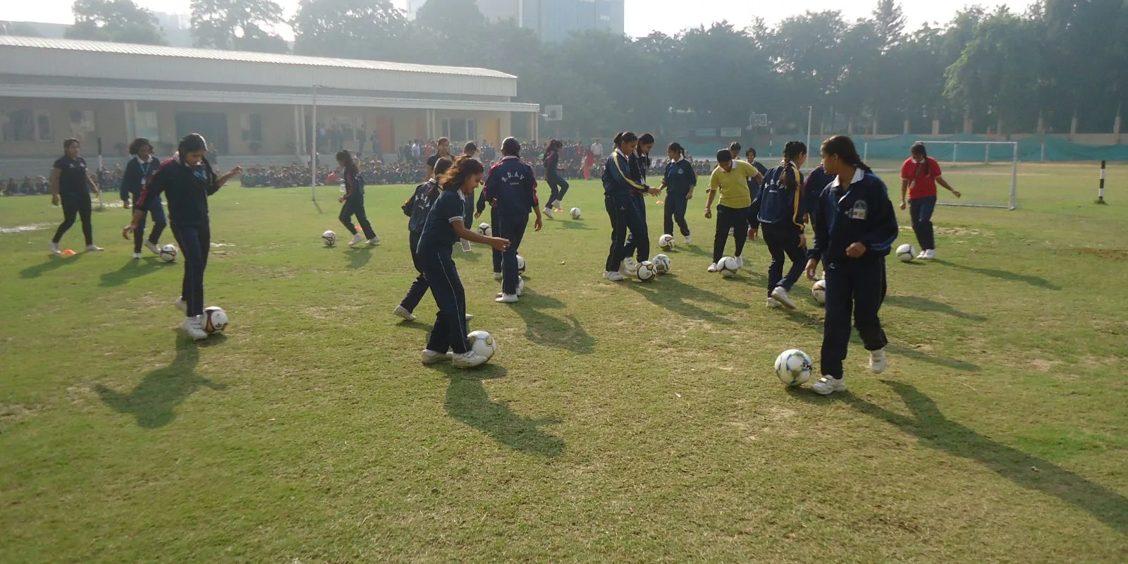 Schoolgirls in tracksuits each dribbling a football across a school ground