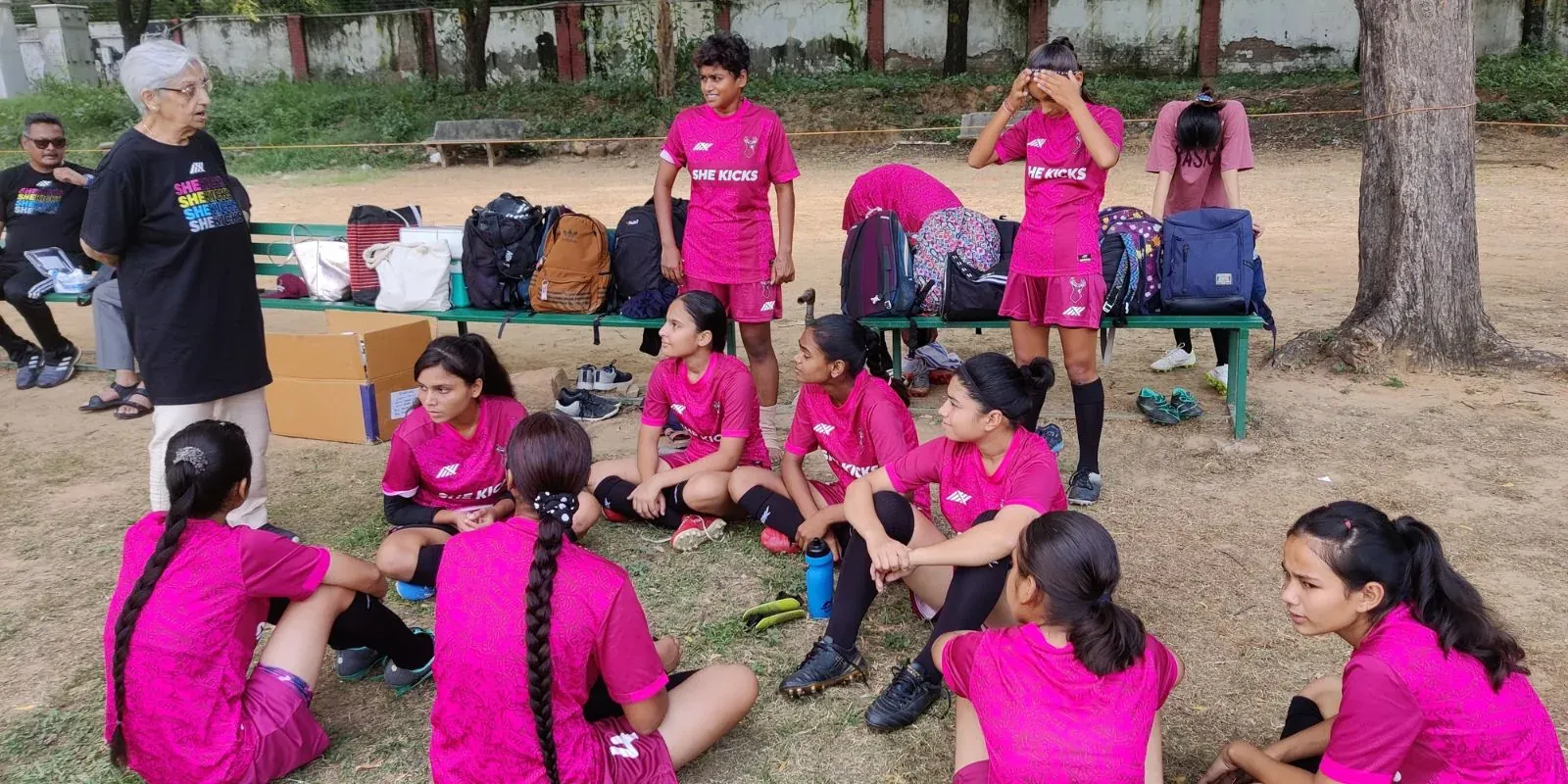 A mentor addressing a seated group of girls in pink kit at a She Kicks Football League event