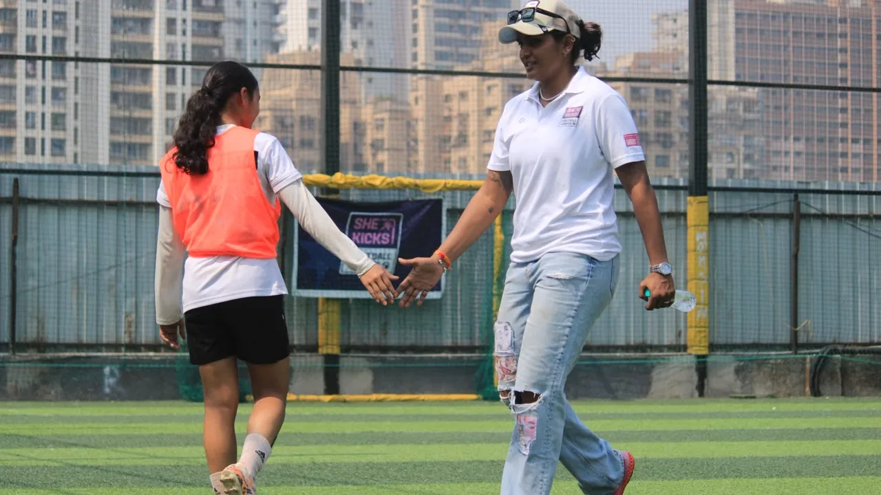 A player in an orange bib and a coach touching hands as they walk across a turf pitch