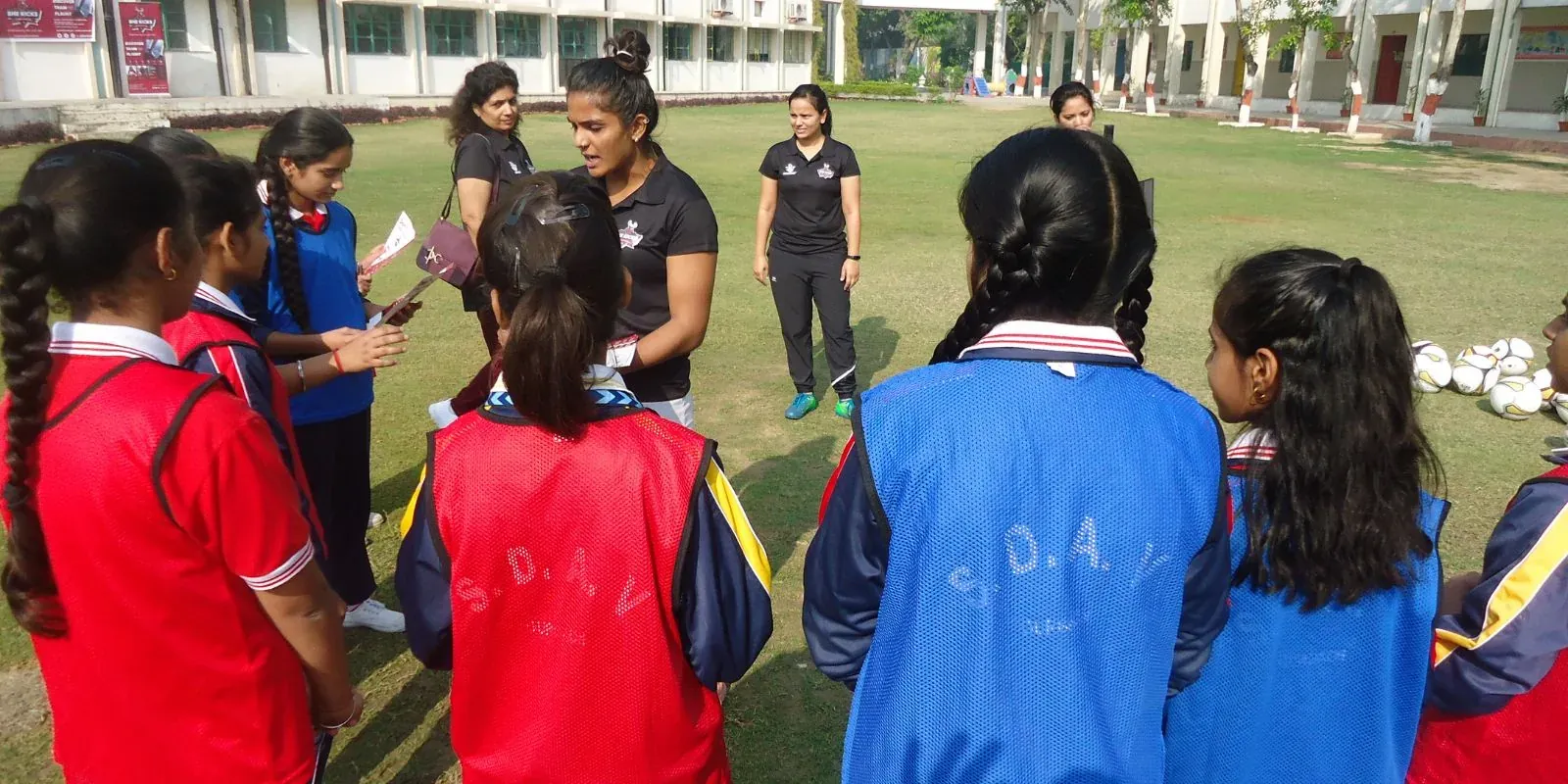 Aditi Chauhan coaching a huddle of schoolgirls in bibs with footballs on a school field